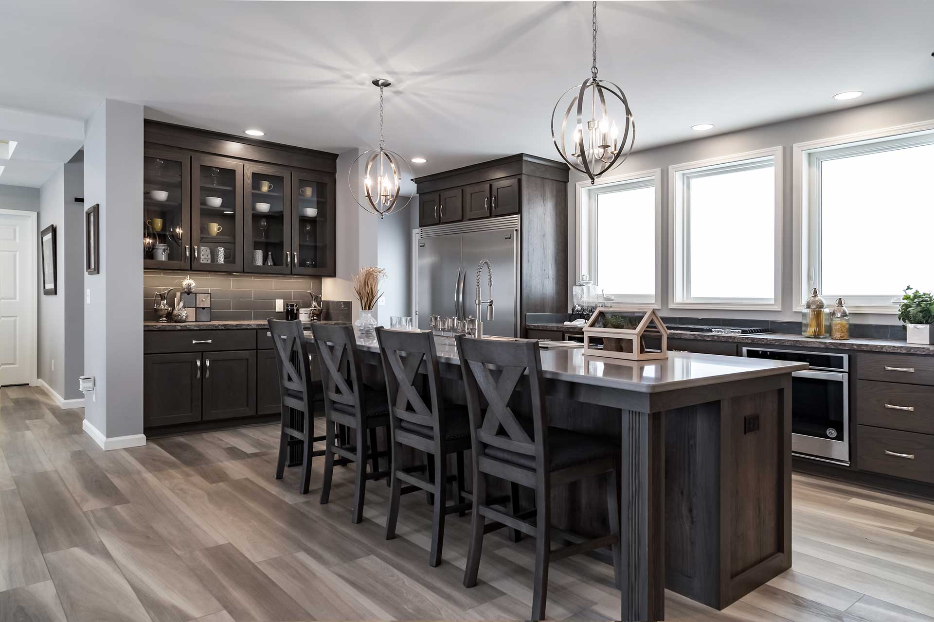 Modern kitchen in the Cavco-Emlenton La Bella Casa manufactured modular home featuring sleek dark walnut toned cabinetry, stainless steel appliances, a coffee station, extra-large center kitchen island with counter-height seating and warm ambient pendant contemporary lighting, luxury kitchen fixtures.